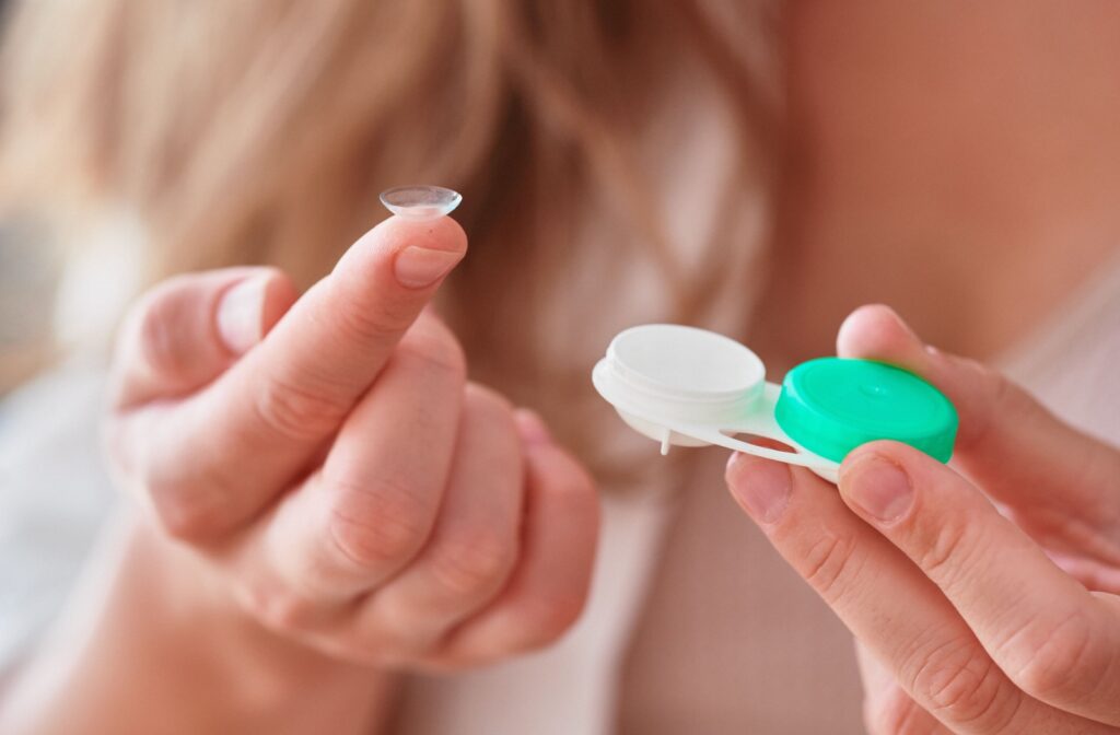 A person holds a contact lens on their fingertip while holding an open contact lens case in the other hand.